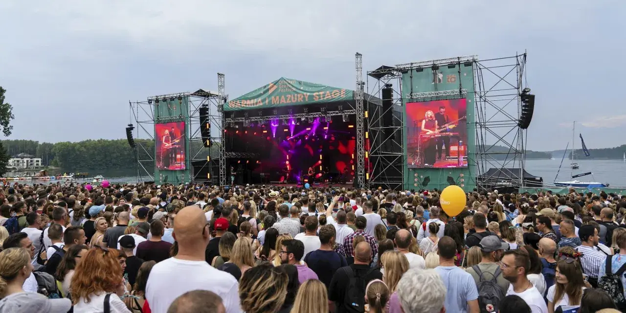 Tłum ludzi na koncercie pod gołym niebem. Na scenie gra zespół, a na ekranach wyświetlany jest green festival line up.