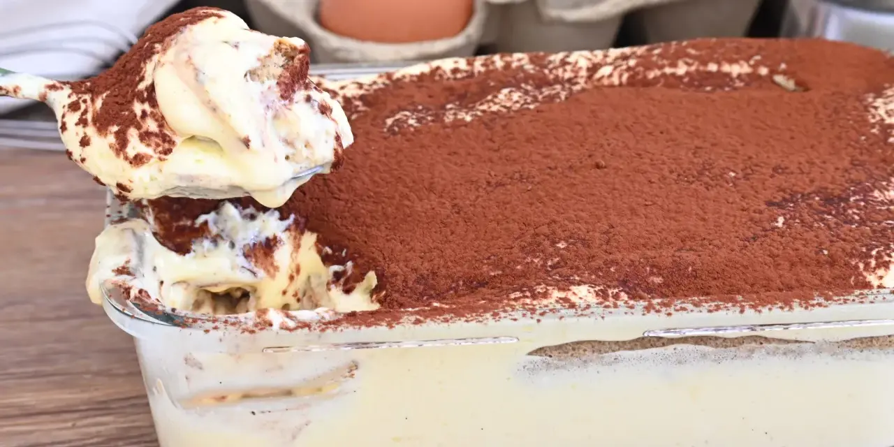 Un cucchiaio preleva una porzione cremosa di tiramisù, con 500 g di mascarpone, cosparso di cacao. Sullo sfondo, uova fresche.