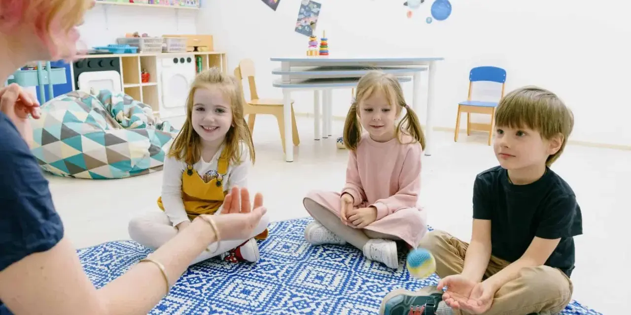 Dzieci siedzą na dywanie, słuchając nauczycielki. To przykład metody dobrego startu, gdzie nauka odbywa się przez zabawę.