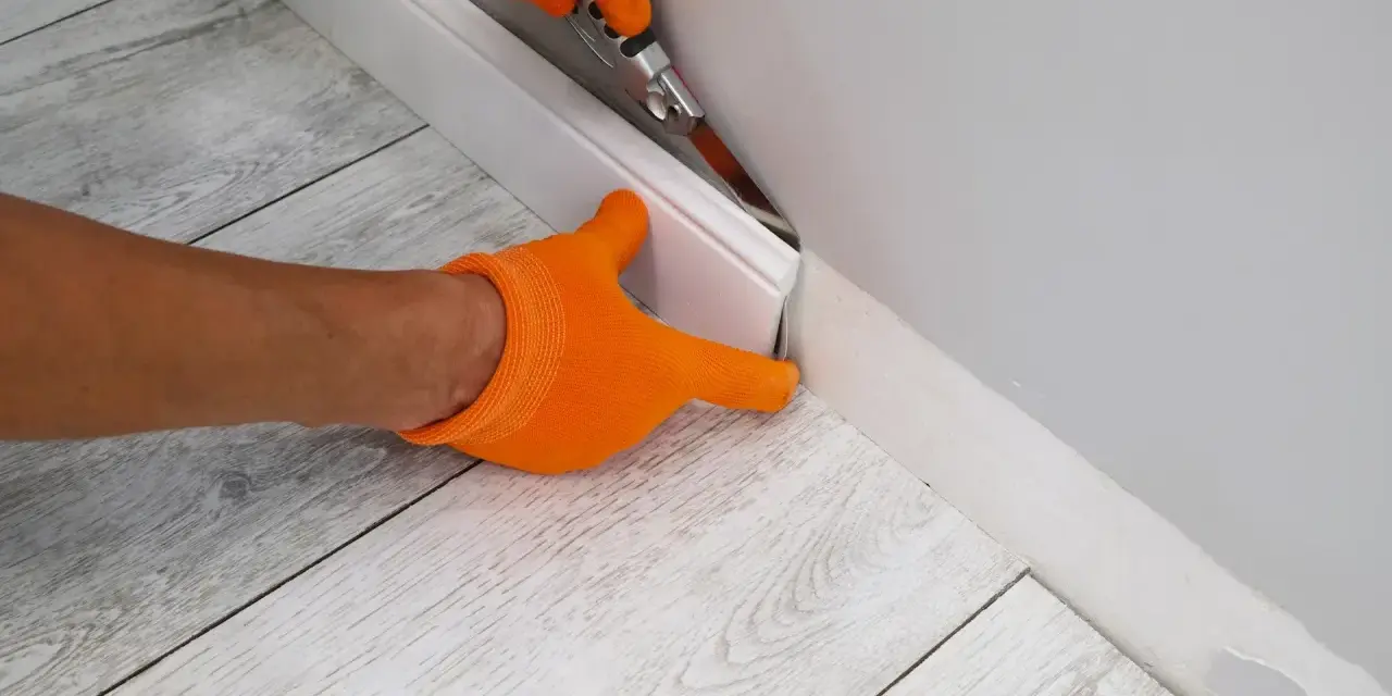 Ręce w pomarańczowych rękawiczkach przycinają listwę przypodłogową, przygotowując ją do wymiany panela podłogowego.