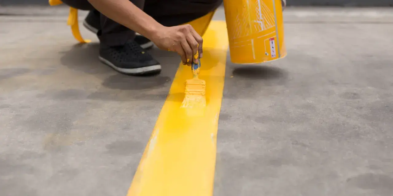 Persona pintando una línea amarilla brillante sobre un suelo de cemento con una brocha.