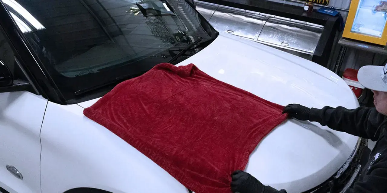 Person in gloves carefully places a plush red towel on a white car hood, demonstrating the best way to wash a car without scratching.
