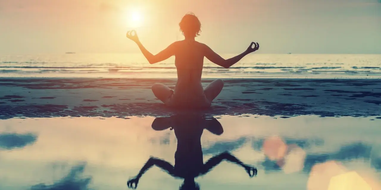 Frau in Lotusposition am Strand bei Sonnenuntergang. Ihre Meditation stärkt das Selbstbewusstsein.