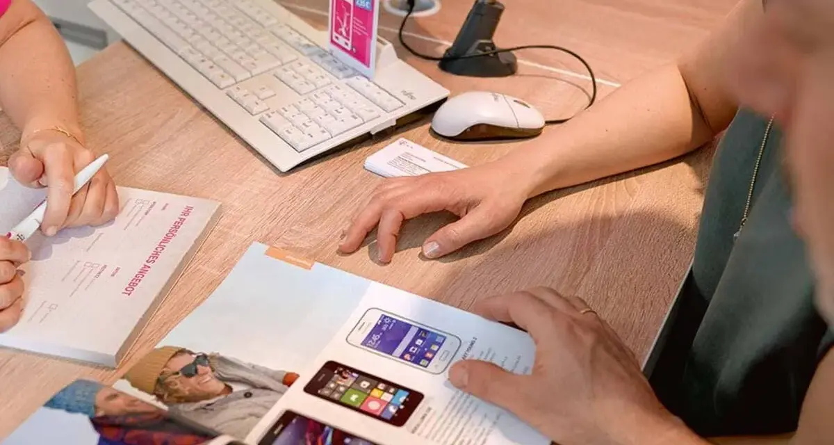 Dwie osoby przeglądają ofertę telefonów komórkowych. Na biurku leży klawiatura, myszka, skaner i katalog z produktami. Jedna z osób pisze coś na formularzu.