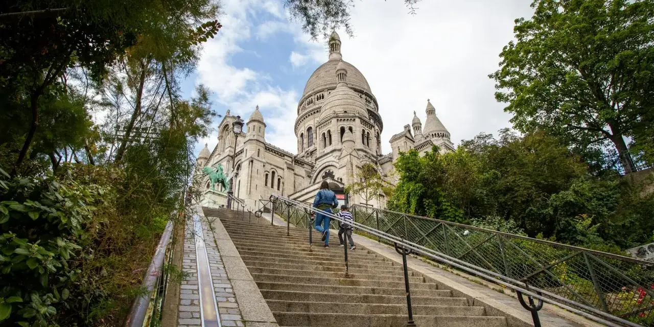 Dwie osoby wspinają się po schodach na wzgórze Montmartre, w tle Bazylika Sacré-Cœur.
