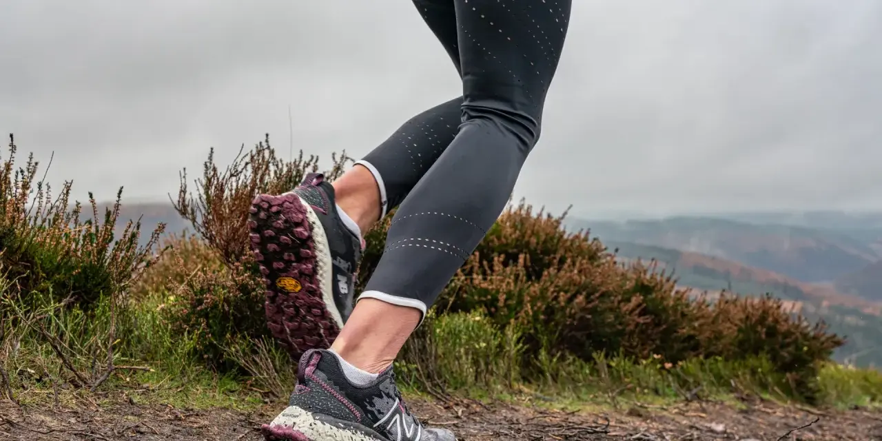 Gambe di una donna che fa trail run su un sentiero fangoso, con erica e colline nebbiose sullo sfondo.