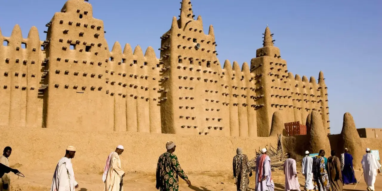 La grande mosquée de Djenné, chef-d'œuvre d'architecture en terre, se dresse sous un ciel bleu. Des hommes en tenues traditionnelles s'approchent, ajoutant vie à ce site historique.