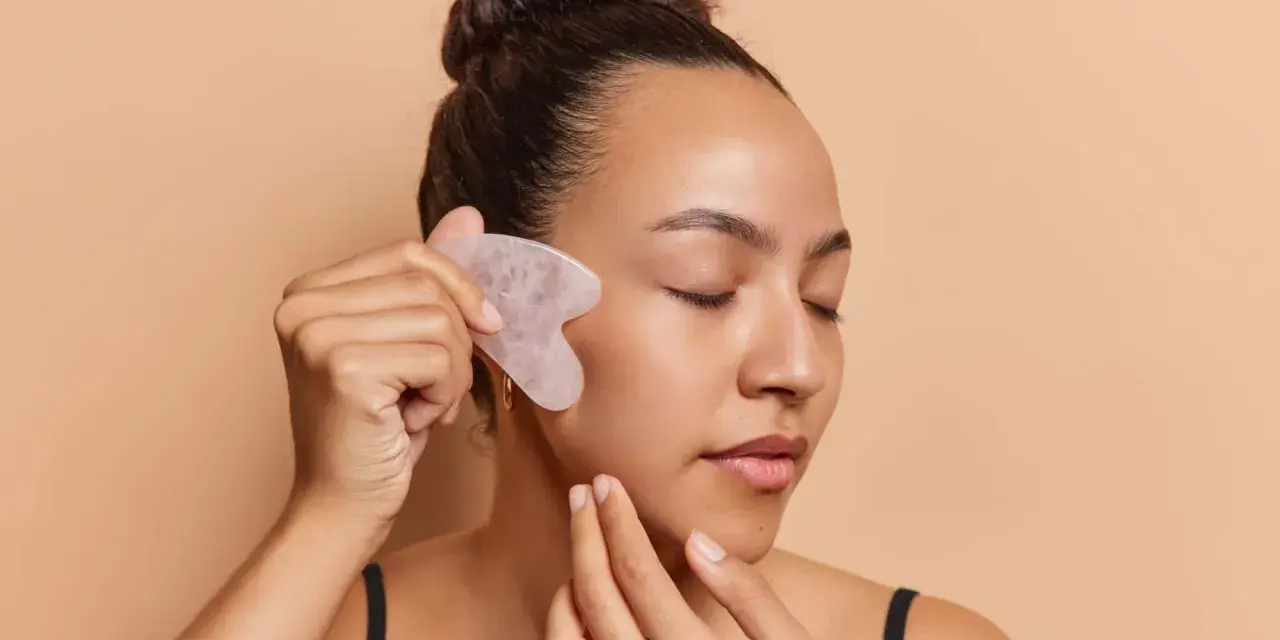 Frau mit geschlossenen Augen nutzt ein Gua Sha Stein zur Gesichtsmassage. Ob morgens oder abends, die Anwendung belebt die Haut.