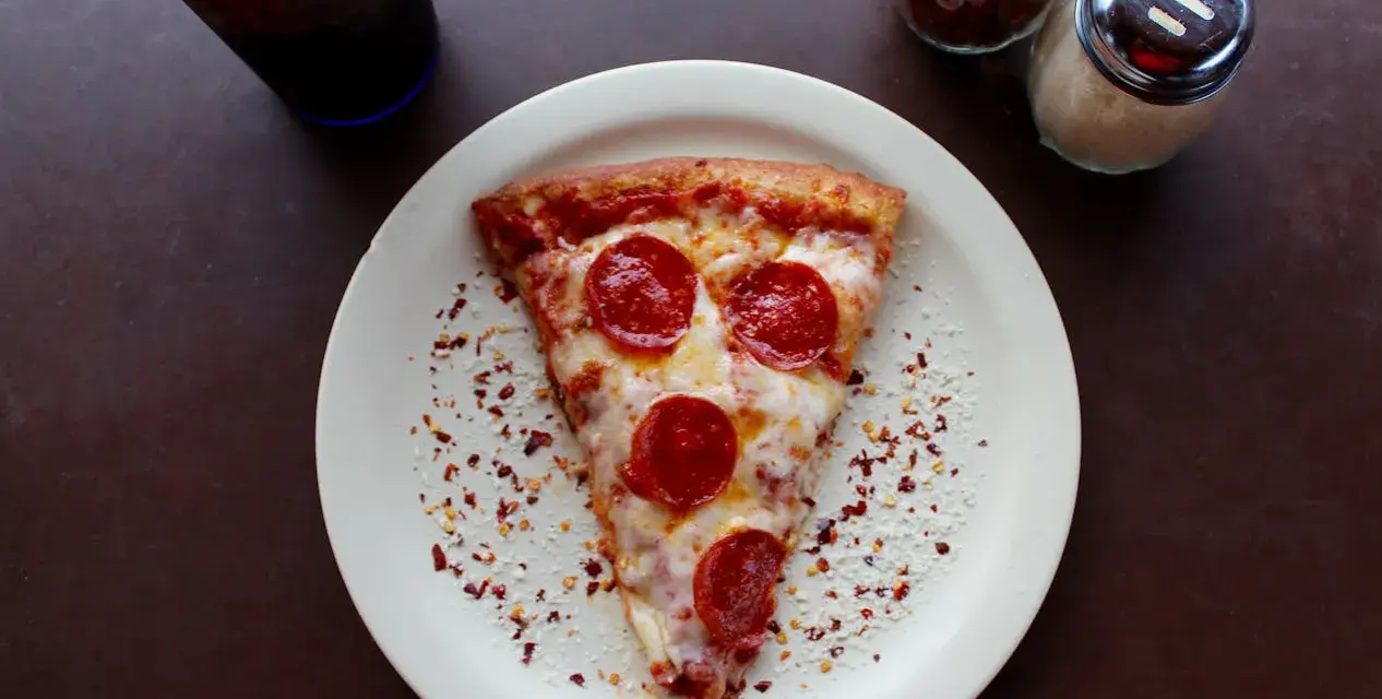Kawałek pizzy pepperoni na talerzu, posypany płatkami chili i parmezanem. Idealny na szybki lunch, choć warto pamiętać o jego kcal.