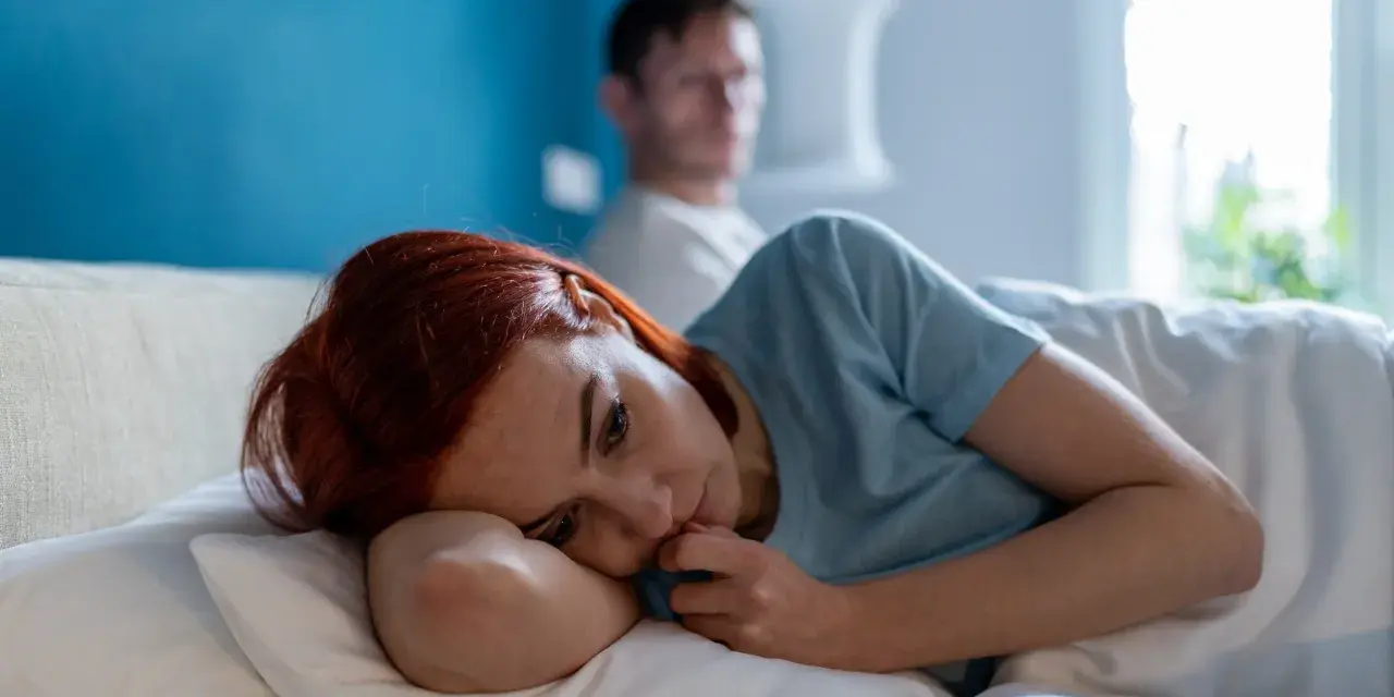 Zamyślona kobieta z rudymi włosami leży w łóżku, zastanawiając się: "czy jestem toksyczna?". W tle niewyraźny mężczyzna.
