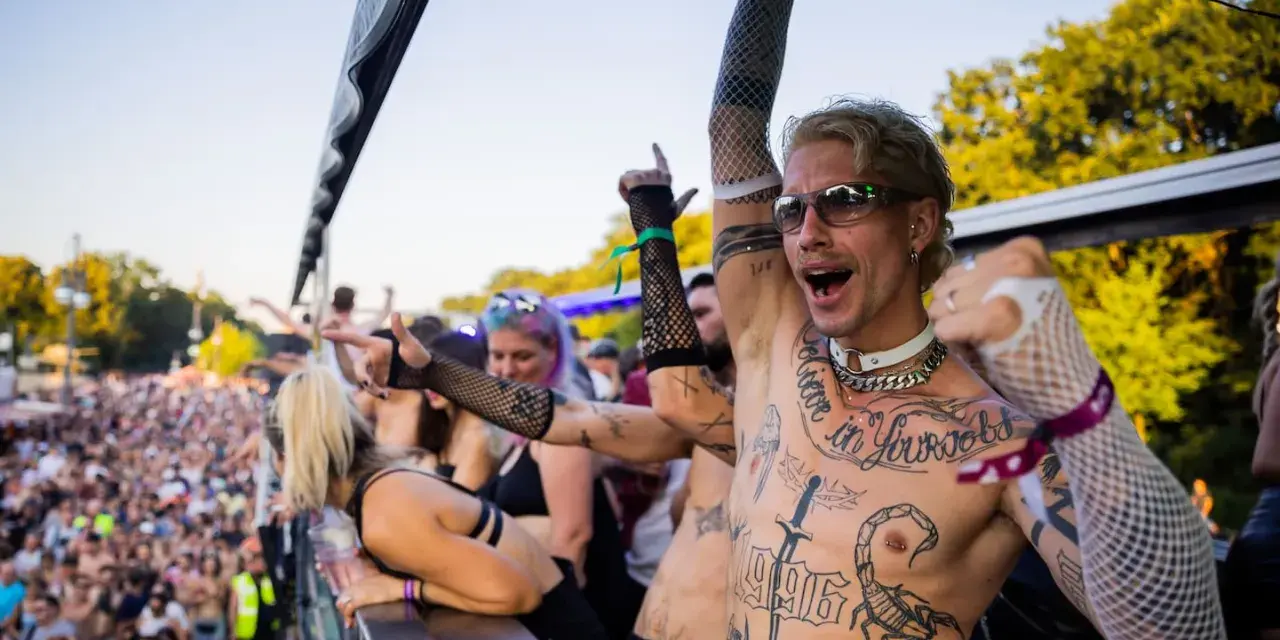 Feiernde Menschenmenge bei einem Festival, die zu **techno music** tanzt. Ein Mann mit Tattoos und Sonnenbrille jubelt.