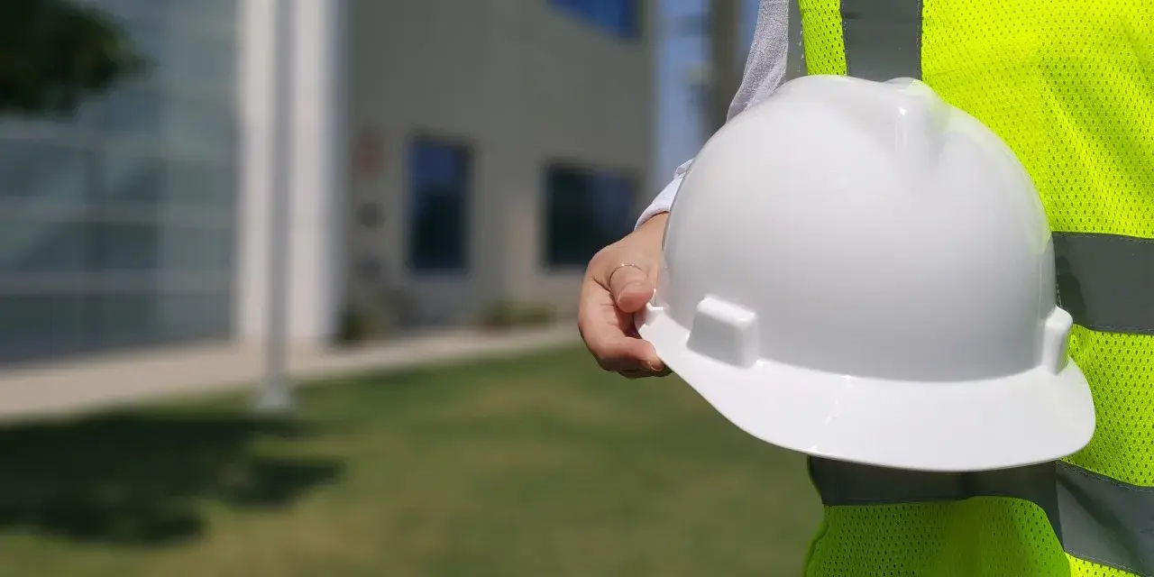 Biały kask w dłoni pracownika na budowie, obok jaskrawożółtej kamizelki. Różne kolory kasków na budowie zapewniają bezpieczeństwo.