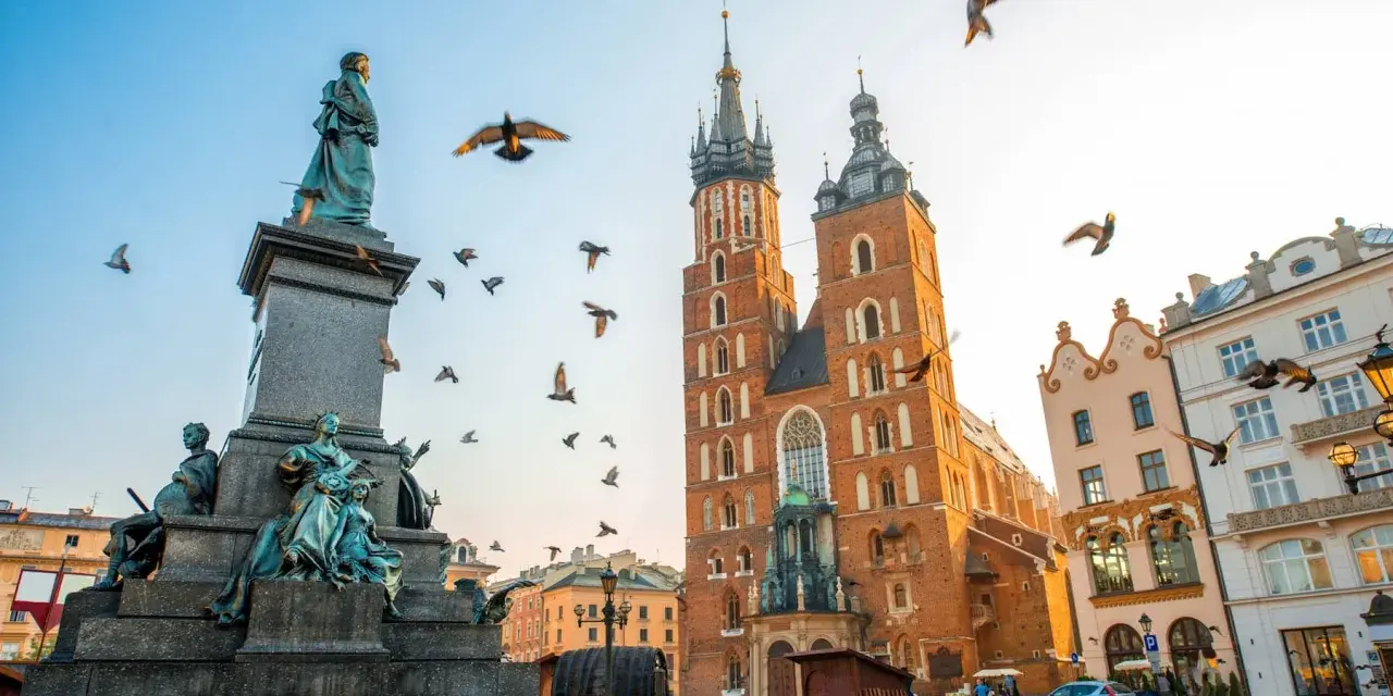 A monument and St. Mary's Basilica dominate this Krakow travel scene. Birds fly around the historic architecture under a clear sky.