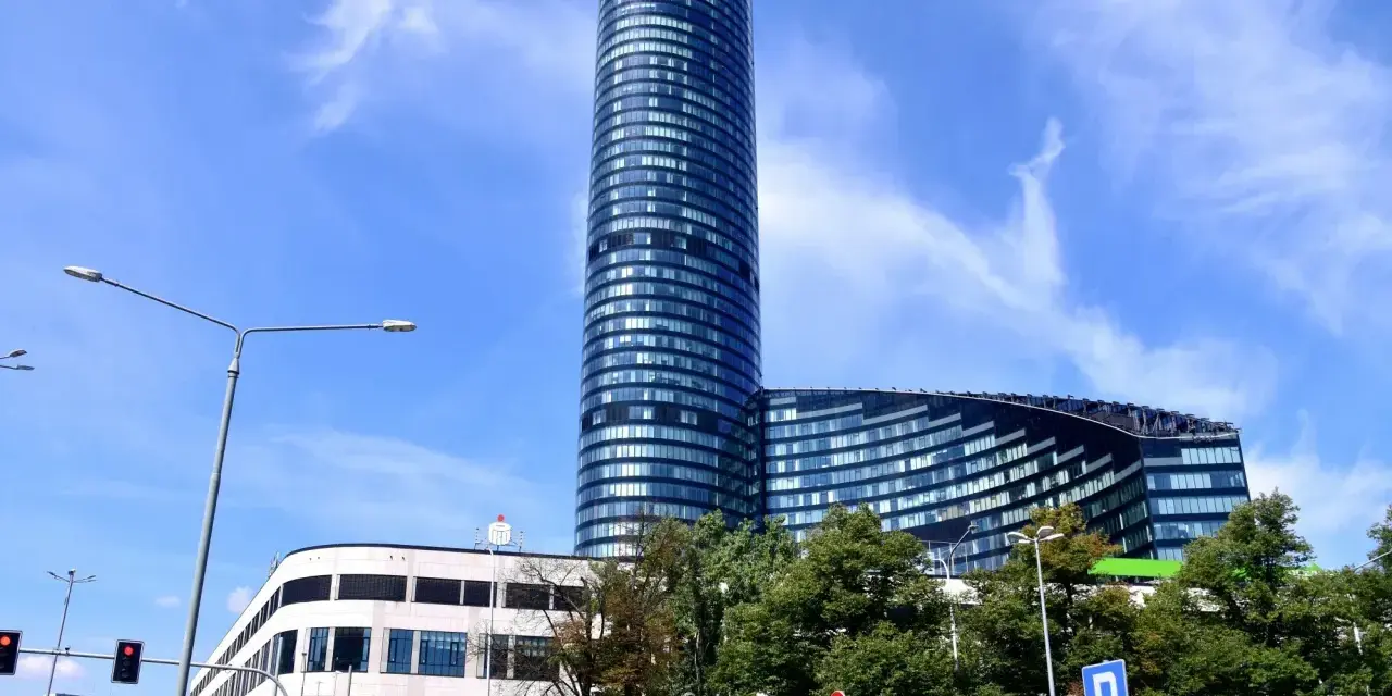 Wysoki, szklany budynek Sky Tower we Wrocławiu, z pewnością ma wiele pięter. Widać też niższy budynek i drzewa.