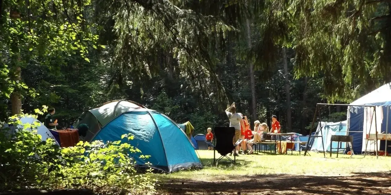 Słoneczny dzień na kempingu. Ludzie siedzą przy stole, rozmawiają, a obok rozstawione są namioty.