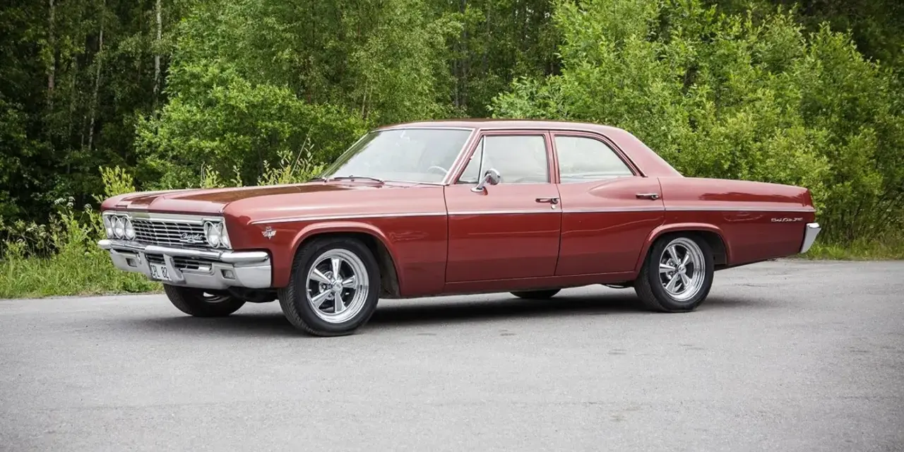 Röd Chevrolet Impala från 1965 med en tung cut, parkerad vid en skogsdunge.
