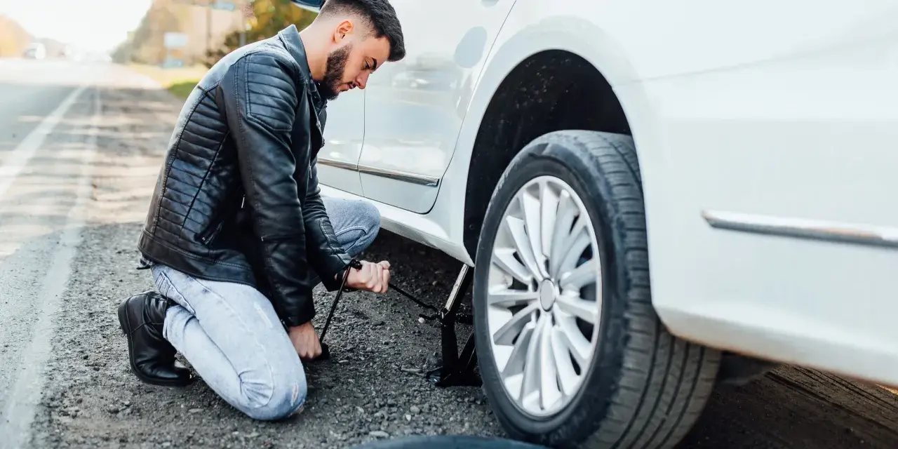 Mężczyzna klęczy przy białym samochodzie, używając podnośnika, by podnieść auto bez kanału. Obok leży zapasowa opona.