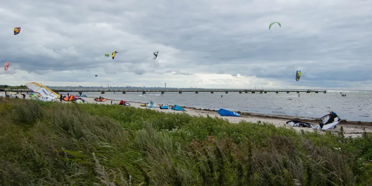 Kitesurfare förbereder sig vid Habo Ljung Camping. Många kites svävar i luften över en strand med en lång pir i bakgrunden.