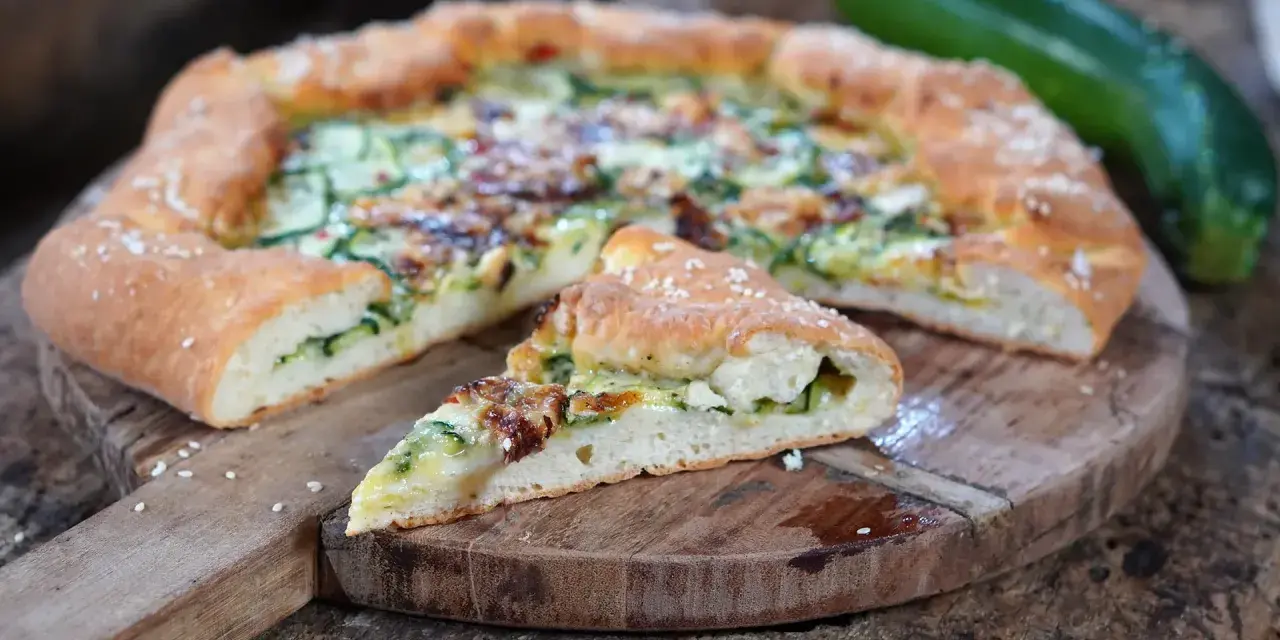 Leckeres Zucchini Pizza Rezept auf einem Holzbrett, mit einem Stück abgetrennt.