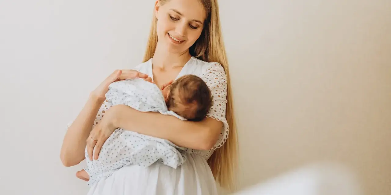 Uśmiechnięta młoda mama tuli noworodka, karmiąc go piersią. Delikatne spojrzenie i biała sukienka podkreślają jej macierzyństwo.