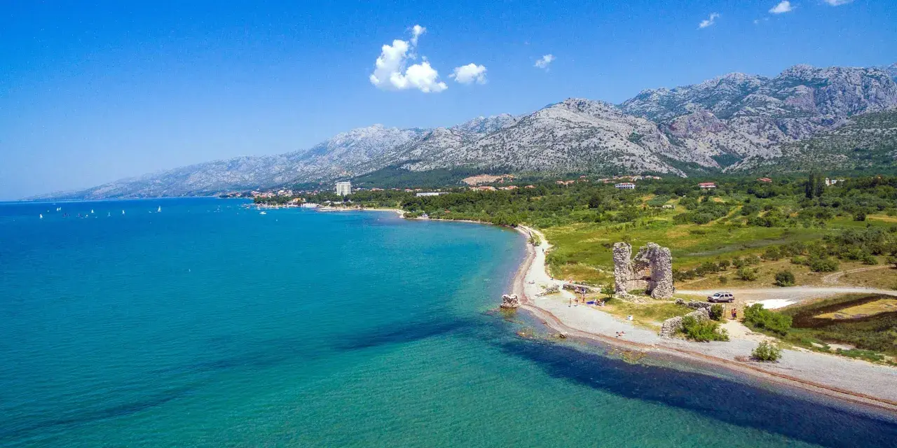 Piękne wybrzeże z górami w tle, idealne na camping Paklenica. Woda kusi turkusowym kolorem, a na plaży widać ruiny starej twierdzy.