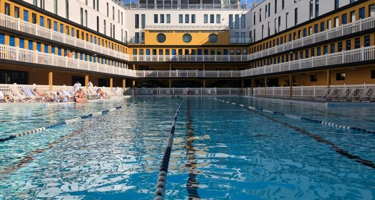 Piscine art déco parisienne, avec ses bâtiments jaunes et blancs, ses balcons et ses transats. L'eau bleue invite à la baignade.