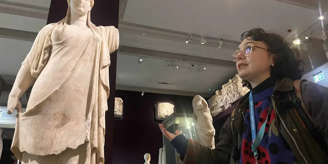 A woman with a museum pass for Turkey gestures towards a marble statue of a woman in a museum.