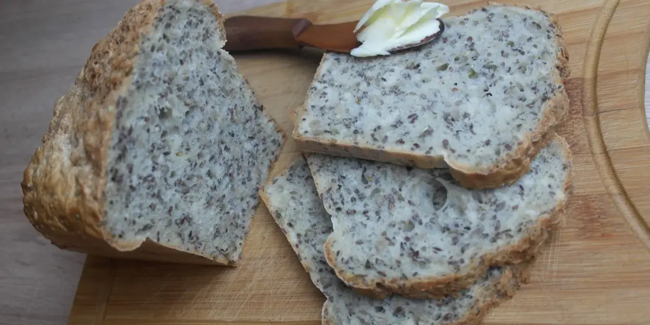 Chleb na proszku do pieczenia, bogaty w ziarna, pokrojony na kromki. Na jednej z nich leży masło na drewnianej łopatce.