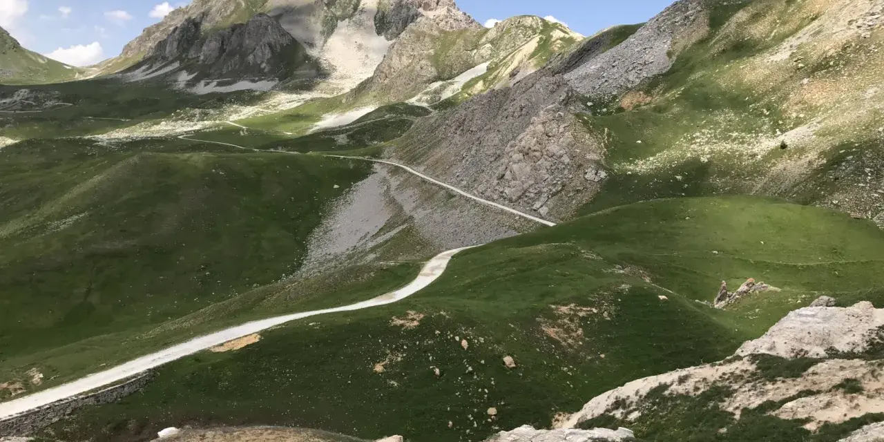 Strada bianca serpeggia tra verdi colline e rocce, verso un monte imponente. Un'avventura in **moto** su una **colle fauniera** panoramica.