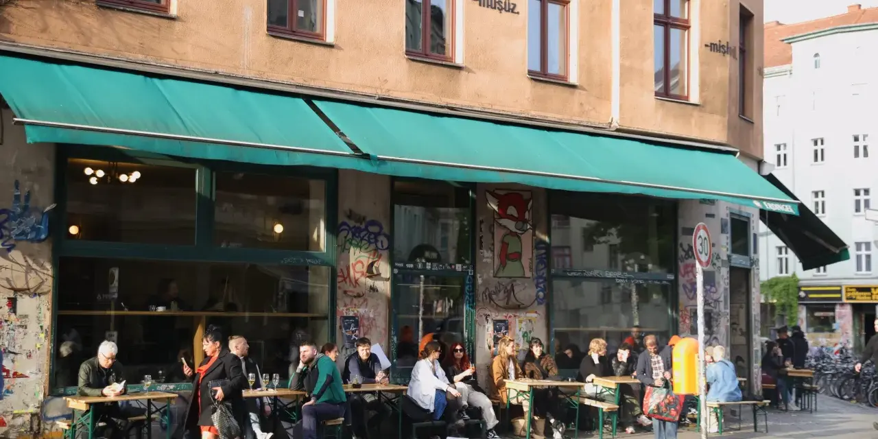 Menschen sitzen an Tischen vor einem Café in Berlin. Die Fassade ist mit Graffiti verziert und trägt die Aufschrift "dorle berlin".