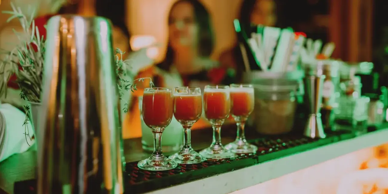 Cztery kieliszki z czerwonym drinkiem stoją na barze, gotowe do podania na weselu. W tle widać gości i dekoracje.