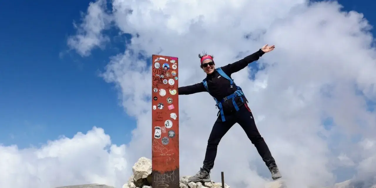 Donna esulta in cima al Monte Amaro dopo un trekking. Cielo azzurro e nuvole bianche.