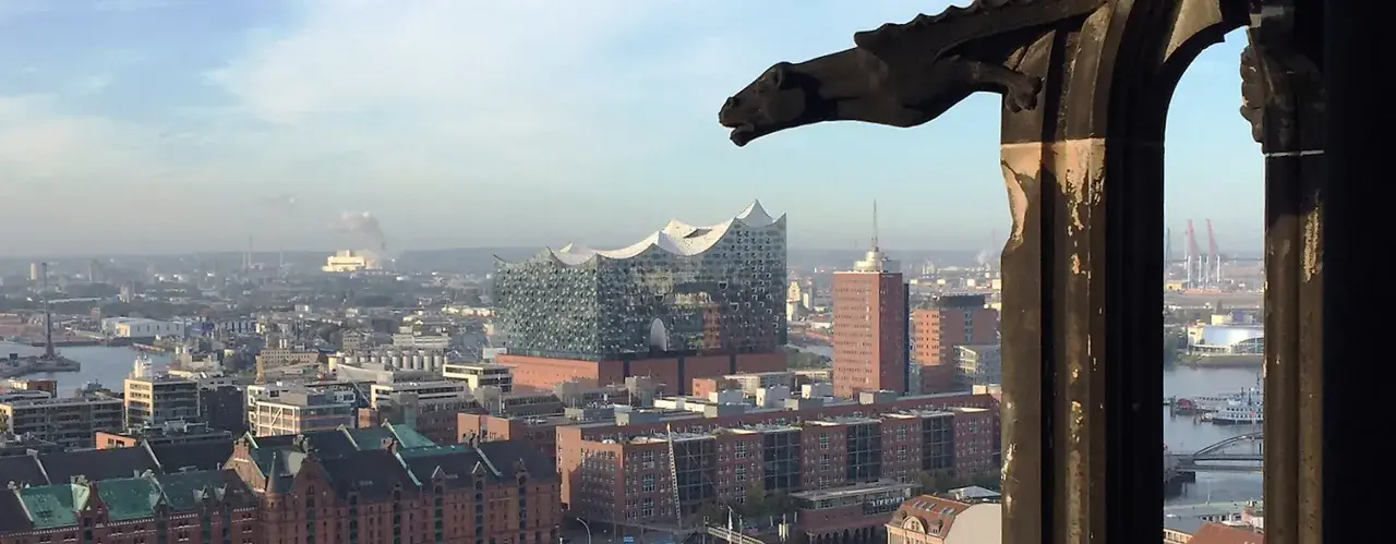 Blick über Hamburg mit der Elbphilharmonie, gesehen durch ein Fenster des Mahnmals St. Nikolai. Ein Wasserspeier wacht über die Stadt.