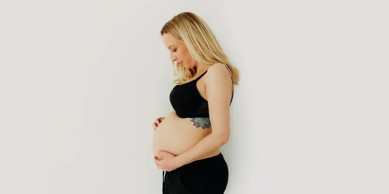 Kobieta w czarnym biustonoszu i spodenkach delikatnie obejmuje swój brzuch w 3 miesiącu ciąży.