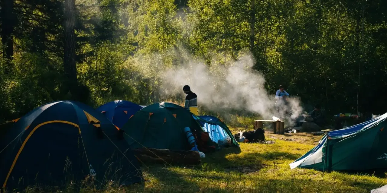 Słoneczny poranek na polu biwakowym. Kilka namiotów rozbitych wśród drzew, unosi się dym z ogniska.