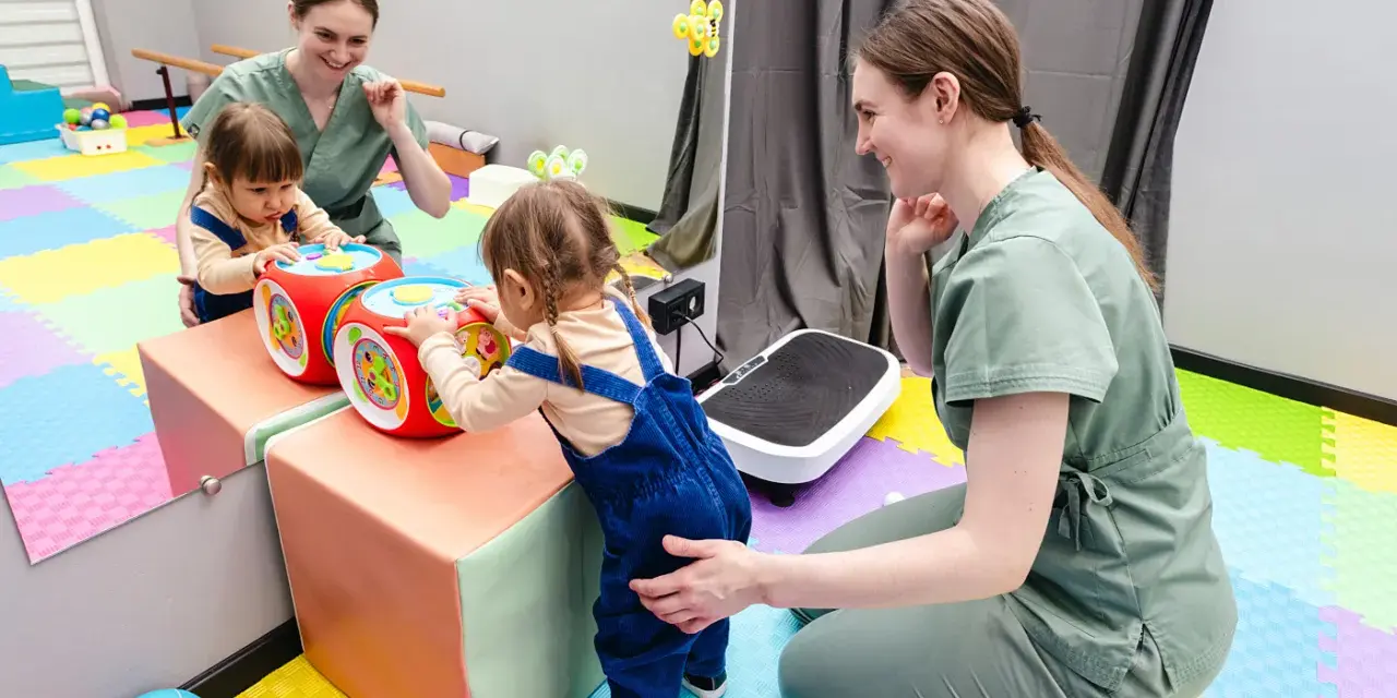 Dwie małe dziewczynki bawią się interaktywną kostką, a fizjoterapeutka wspiera jedną z nich. To może być część terapii wwr co to.