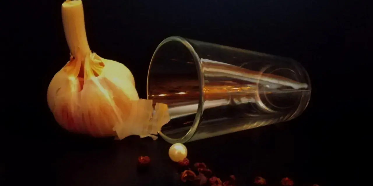 Photographie nature morte : une tête d'ail, un verre renversé, une perle et des baies de poivre rouge sur fond sombre.