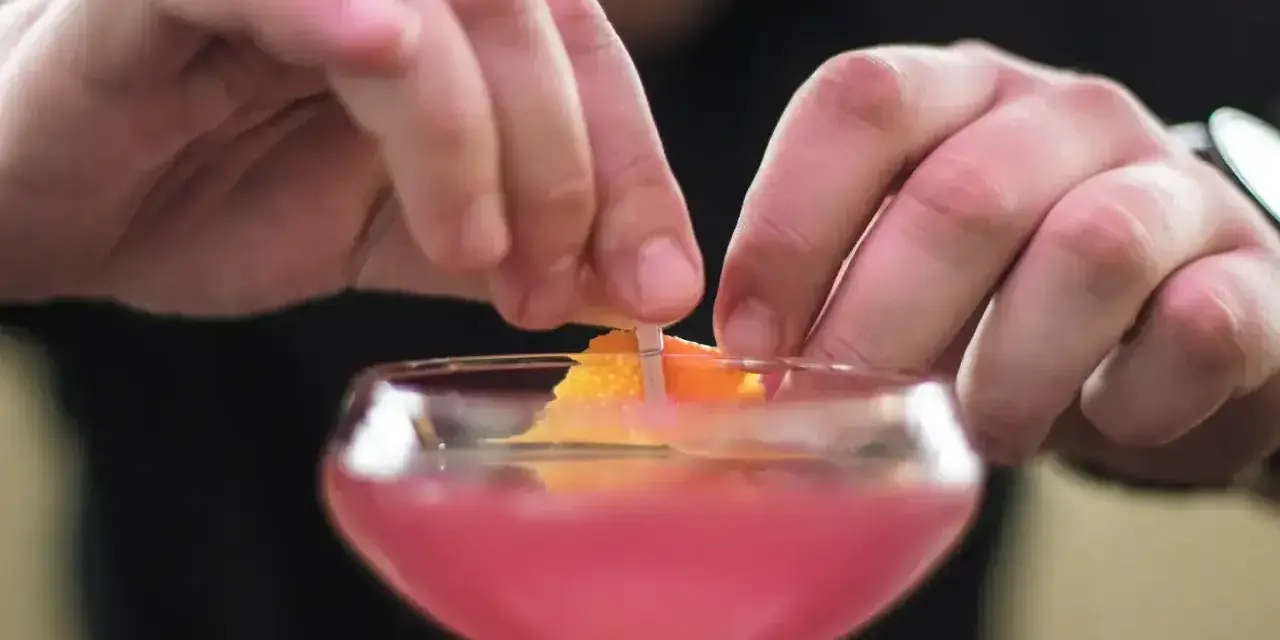 Dłonie barmana ozdabiają drinka skórką pomarańczy. To klasyczny cosmopolitan, gotowy na wieczór w wielkim mieście.