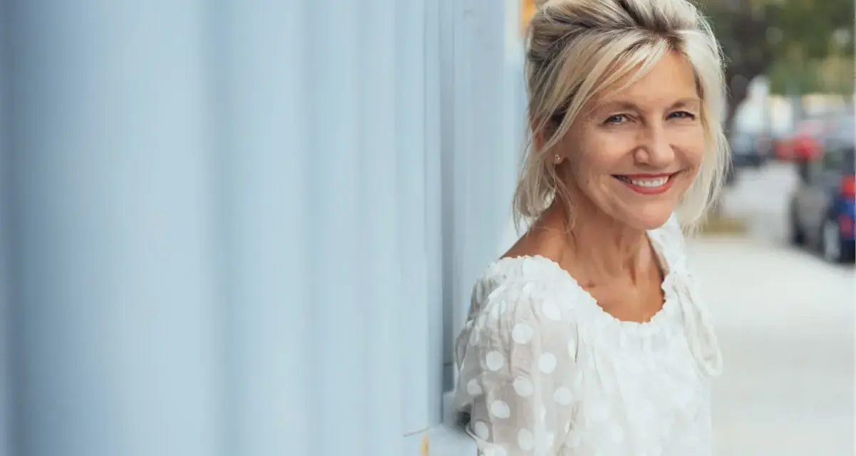 Donna sorridente appoggiata a un muro blu. Potrebbe beneficiare di acido folico in menopausa per il suo benessere.