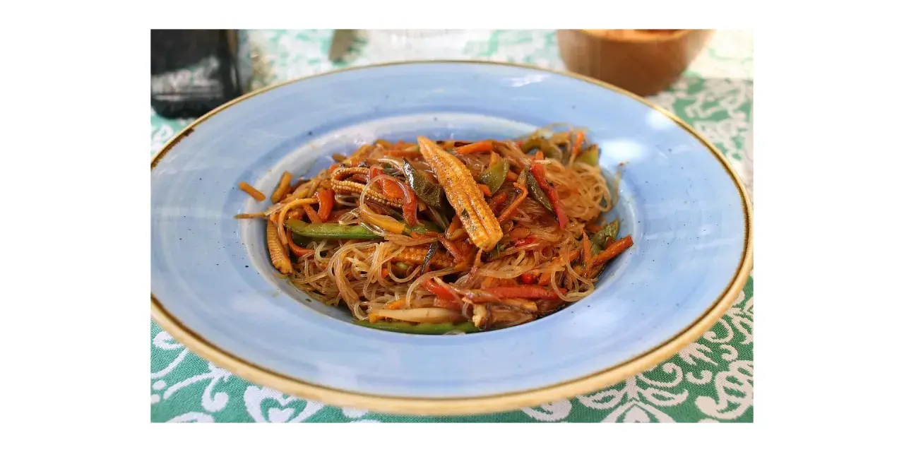 Un piatto di spaghetti di riso con verdure colorate, tra cui peperoni, carote e pannocchiette, condito con una salsa saporita.