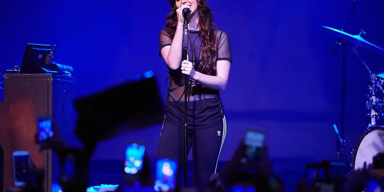 Lana Del Rey, w czarnej, prześwitującej bluzce i sportowych spodniach, śpiewa na scenie. Fani z telefonami tworzą tłum. To jej lana del rey opener.