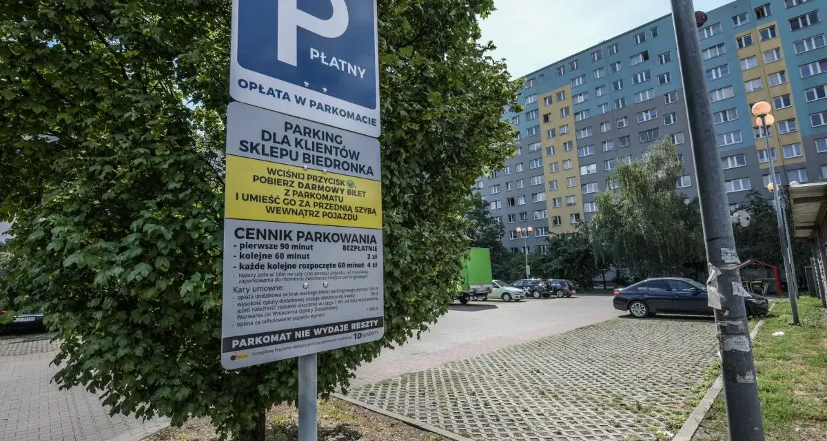 Płatny parking dla klientów Biedronki. Obok drzewa stoi znak informujący o cenniku i zasadach parkowania.