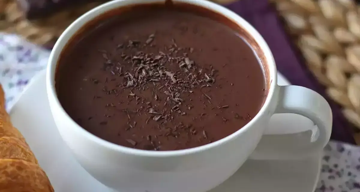 En vit mugg med varm choklad, toppad med chokladspån. En croissant ligger bredvid.