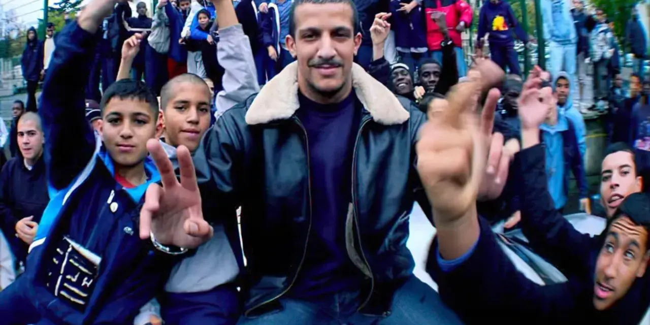 Un groupe de jeunes pose pour une photo, certains font des signes de paix. L'ambiance est celle du rap francais.