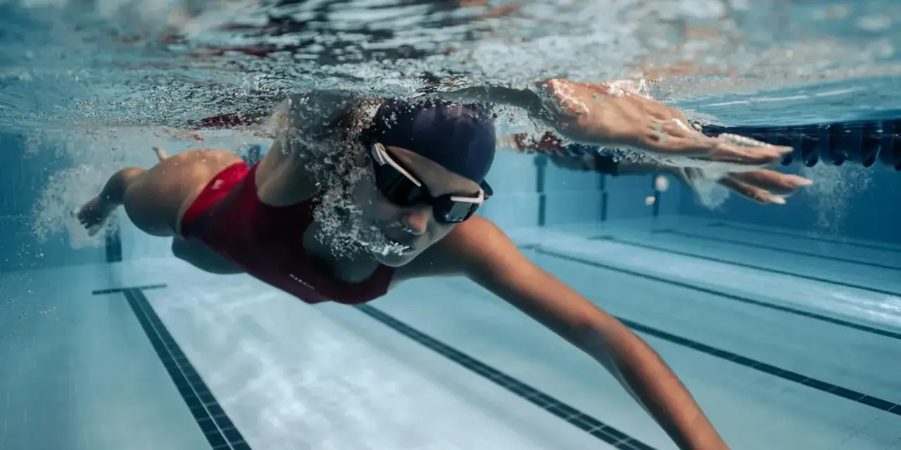Une femme en maillot de bain rouge nage en crawl, lunettes de natation sur le nez. Sa **natation transformation physique femme** est visible sous l'eau.