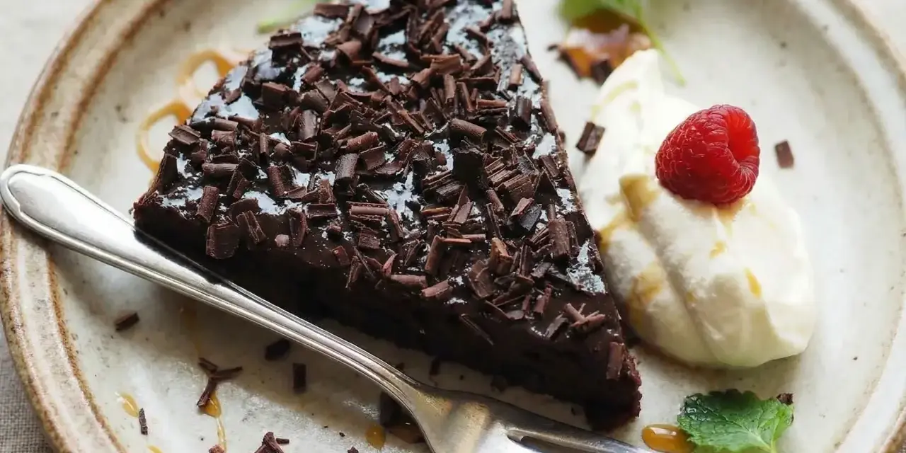 En bit glutenfri kladdkaka toppad med chokladspån, serverad med vispad grädde, hallon och mynta.