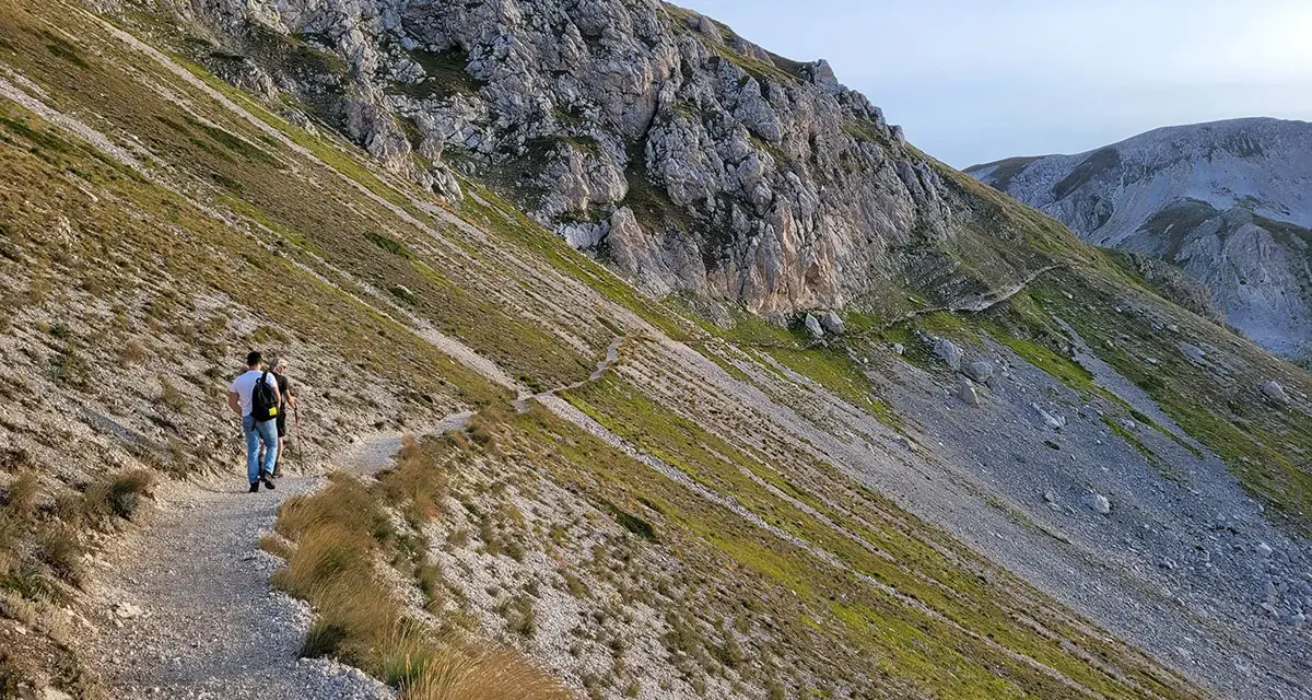 Due escursionisti percorrono un sentiero di trekking sul Gran Sasso, tra rocce e vegetazione alpina.