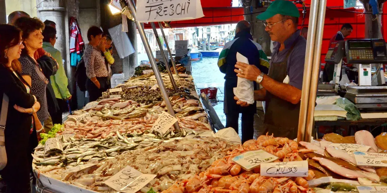 Mercato Venezia: bancarella di pesce fresco con gamberi, calamari e pesciolini. Venditore con cappellino verde.