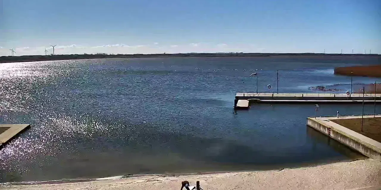 Widok z jeziorsko kamera na żywo. Na piaszczystej plaży dwie osoby, w tle spokojne jezioro, pomost i wiatraki.