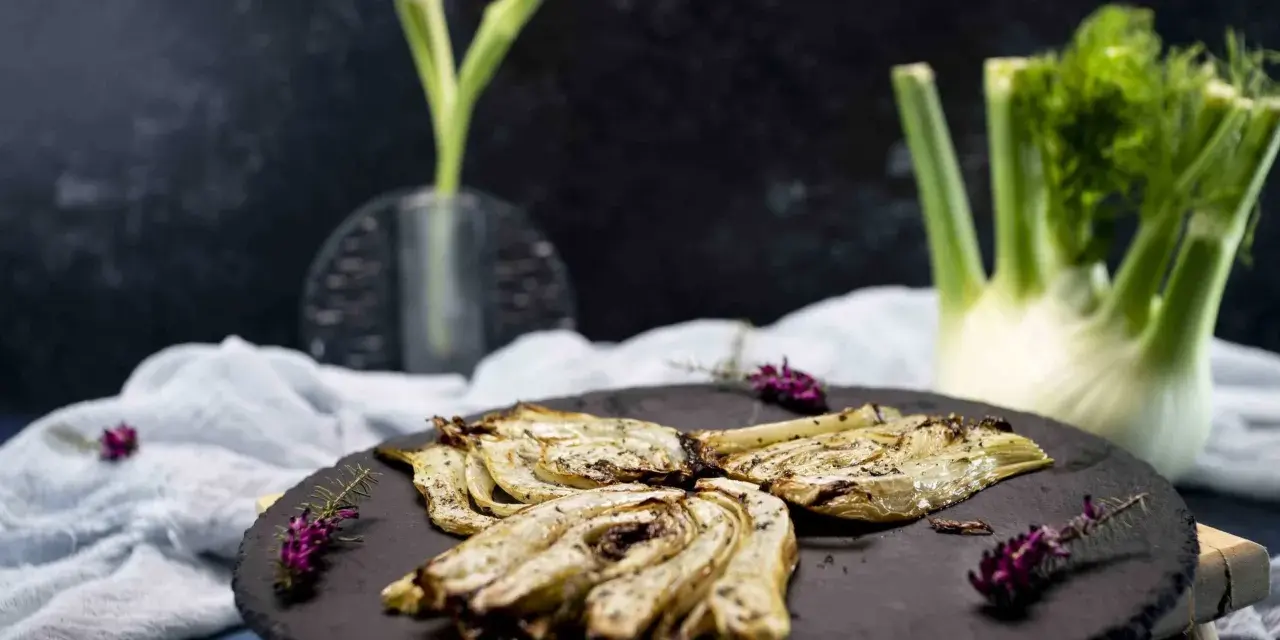 Finocchi croccanti e dorati, cotti nella friggitrice ad aria, serviti su una pietra scura con erbe aromatiche.